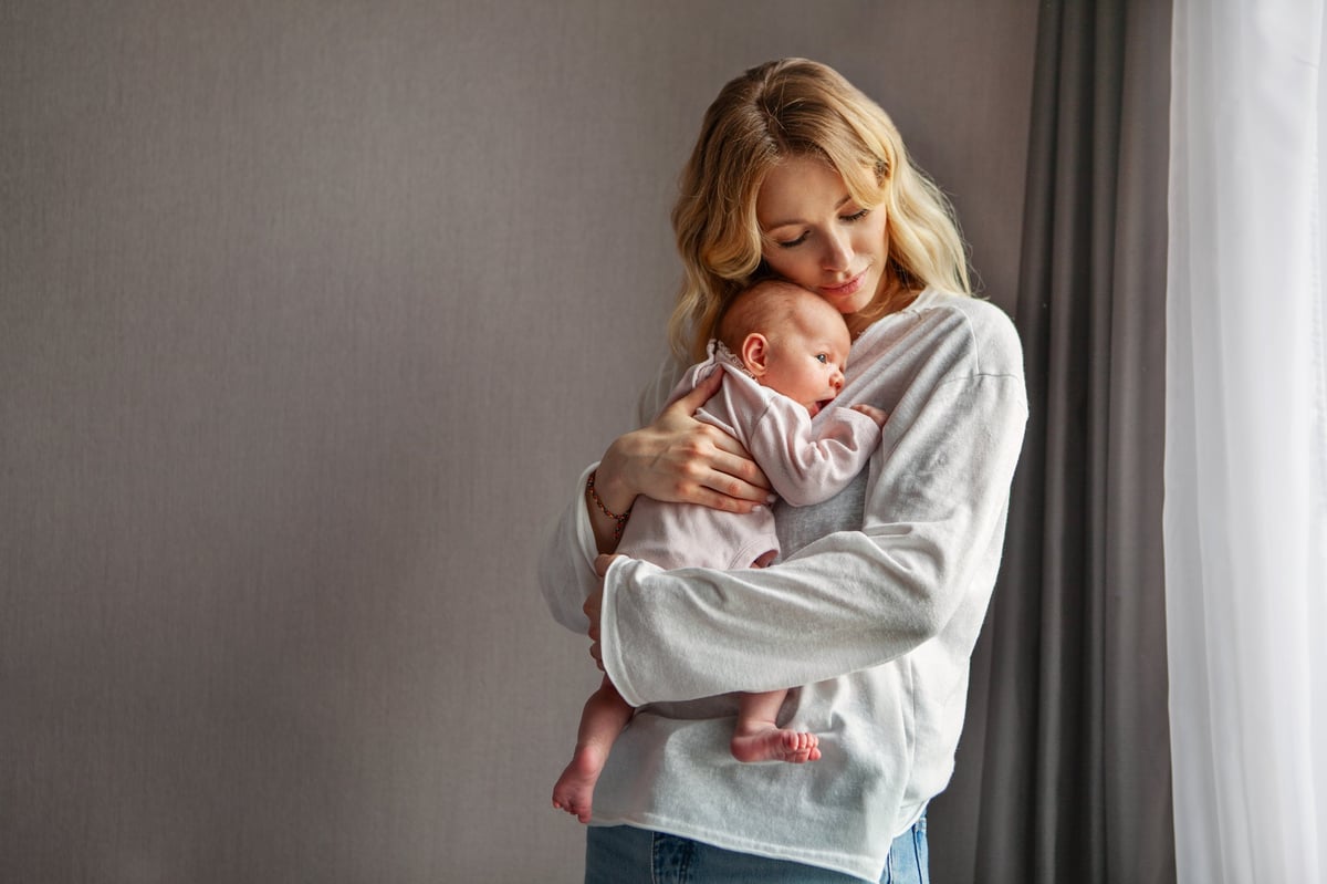 Young mother holding newborn baby near window, tender moment in natural light